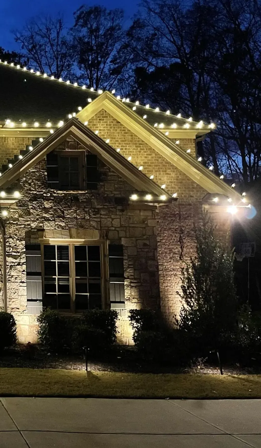 Warm-white Christmas lights along the roofline and gable peaks of a large two-story brick and stone home